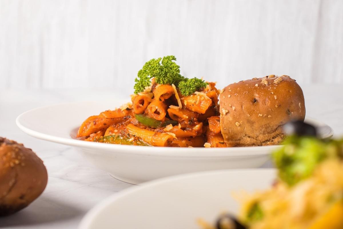 A plate of penne pasta tossed in a rich, red tomato sauce and topped with grated cheese and fresh green parsley. A small, crusty bread roll sits on the edge of the white bowl, set against a light background with other dishes blurred in the foreground. A plate of penne pasta tossed in a rich, red tomato sauce and topped with grated cheese and fresh green parsley. A small, crusty bread roll sits on the edge of the white bowl, set against a light background with other dishes blurred in the foreground.
