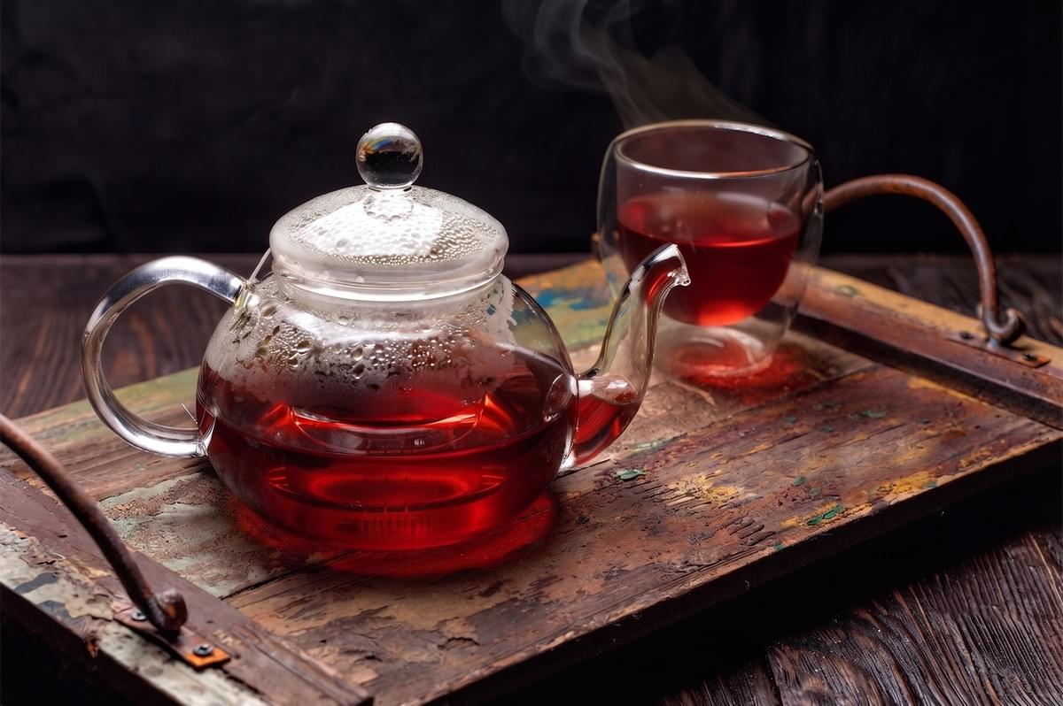 Glass teapot and matching double-walled cup filled with steaming red herbal tea on a rustic wooden tray. Glass teapot and matching double-walled cup filled with steaming red herbal tea on a rustic wooden tray.
