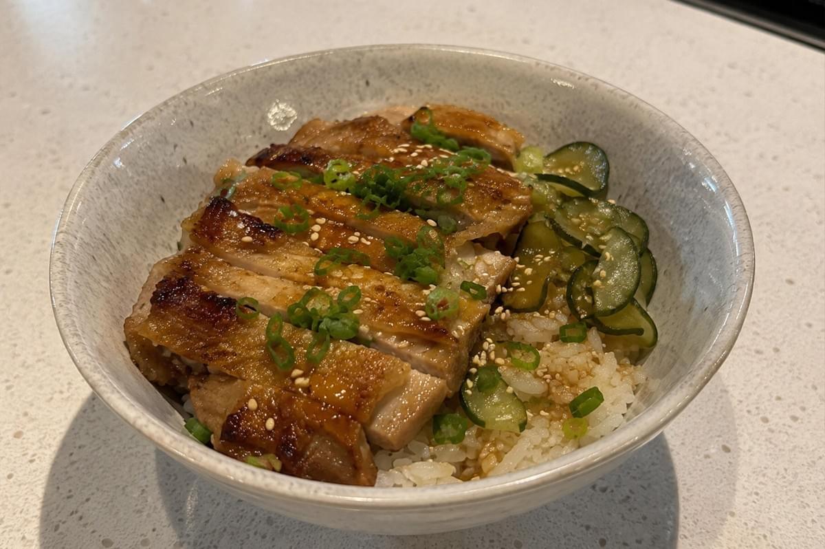 A top-down, slightly angled view of a completed chicken rice bowl served in a textured, off-white ceramic bowl. The dish features succulent, sliced chicken with a caramelized glaze, arranged neatly next to a cluster of vinegared cucumber slices, with the entire meal finished with a garnish of green onions and sesame seeds. A top-down, slightly angled view of a completed chicken rice bowl served in a textured, off-white ceramic bowl. The dish features succulent, sliced chicken with a caramelized glaze, arranged neatly next to a cluster of vinegared cucumber slices, with the entire meal finished with a garnish of green onions and sesame seeds.