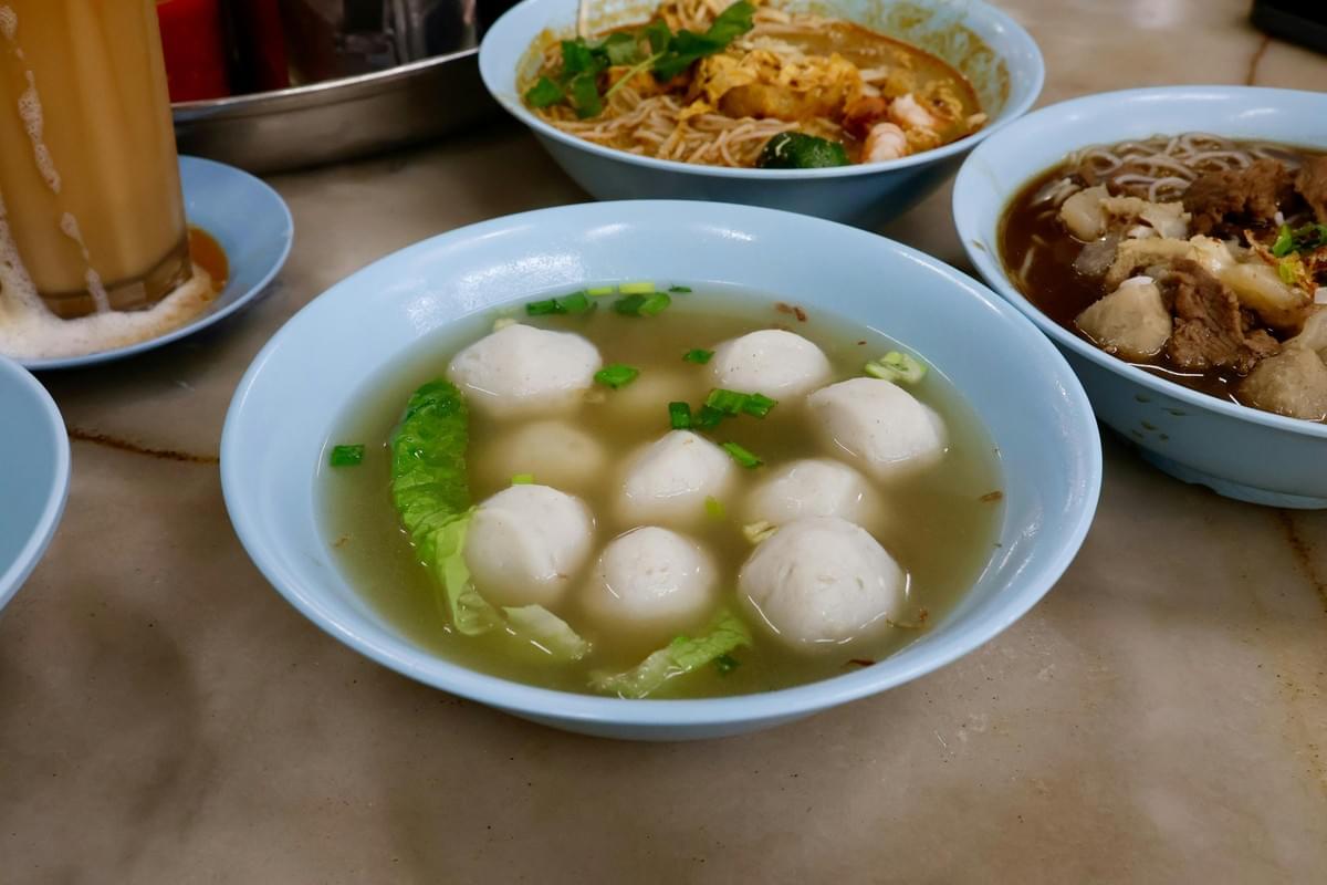 A blue bowl filled with clear soup, white fish balls, scallions, and greens rests in the foreground on a table. In the background, other noodle dishes and a frothy iced milk tea sit nearby. A blue bowl filled with clear soup, white fish balls, scallions, and greens rests in the foreground on a table. In the background, other noodle dishes and a frothy iced milk tea sit nearby.
