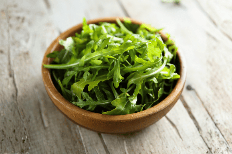 A small, rustic brown bowl filled with crisp, peppery arugula greens sitting on a weathered wooden table. A small, rustic brown bowl filled with crisp, peppery arugula greens sitting on a weathered wooden table.