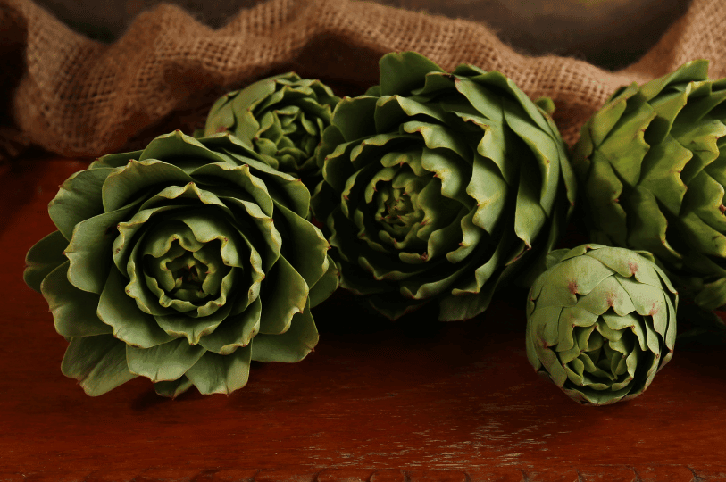 Artistic arrangement of five dark green artichokes on a rustic wooden surface with a burlap backdrop, highlighting their blooming, rose-like shape. Artistic arrangement of five dark green artichokes on a rustic wooden surface with a burlap backdrop, highlighting their blooming, rose-like shape.