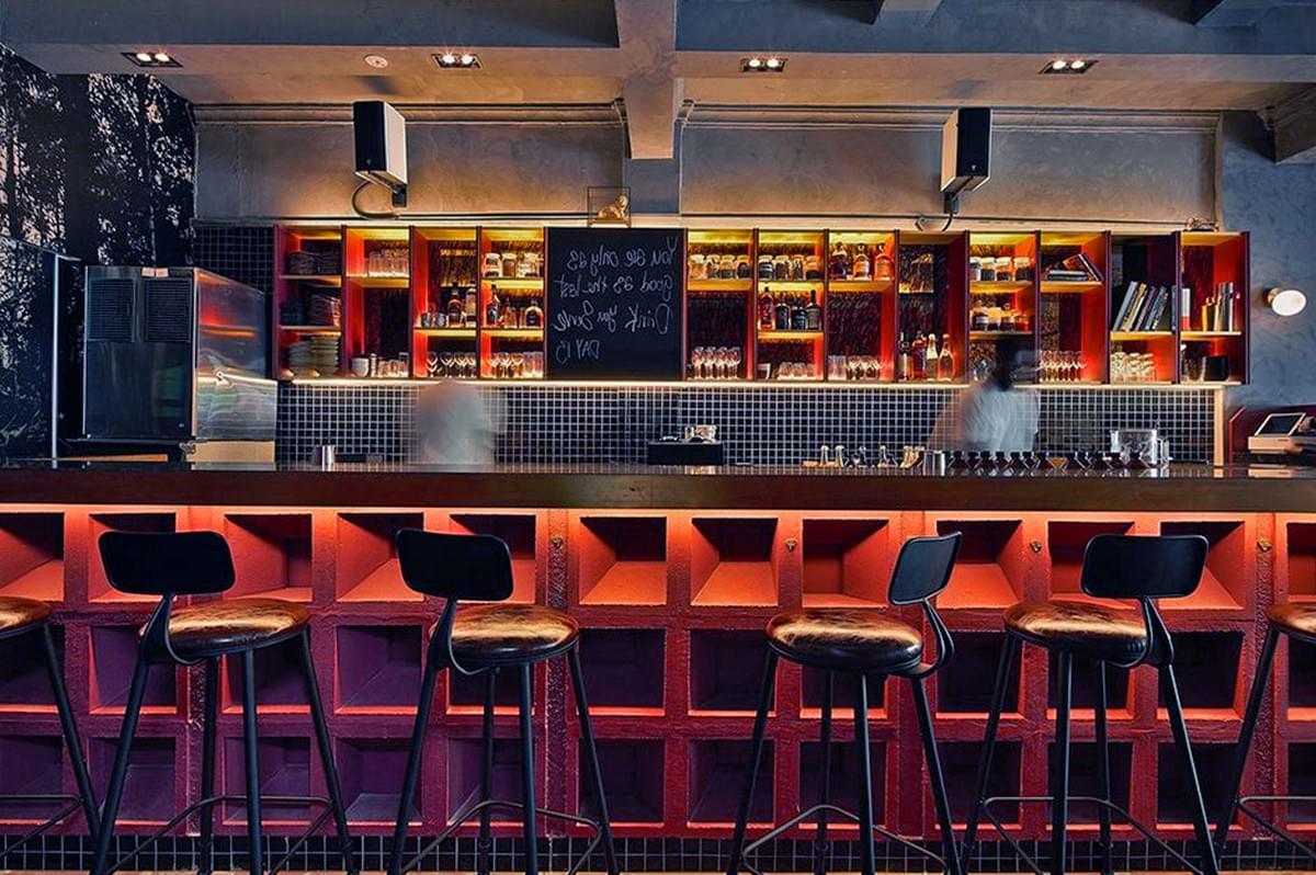 Cozy bar interior with warm lighting. Wooden bar stools in front, shelves filled with bottles, glasses, and a chalkboard. Inviting, vibrant atmosphere. Cozy bar interior with warm lighting. Wooden bar stools in front, shelves filled with bottles, glasses, and a chalkboard. Inviting, vibrant atmosphere.