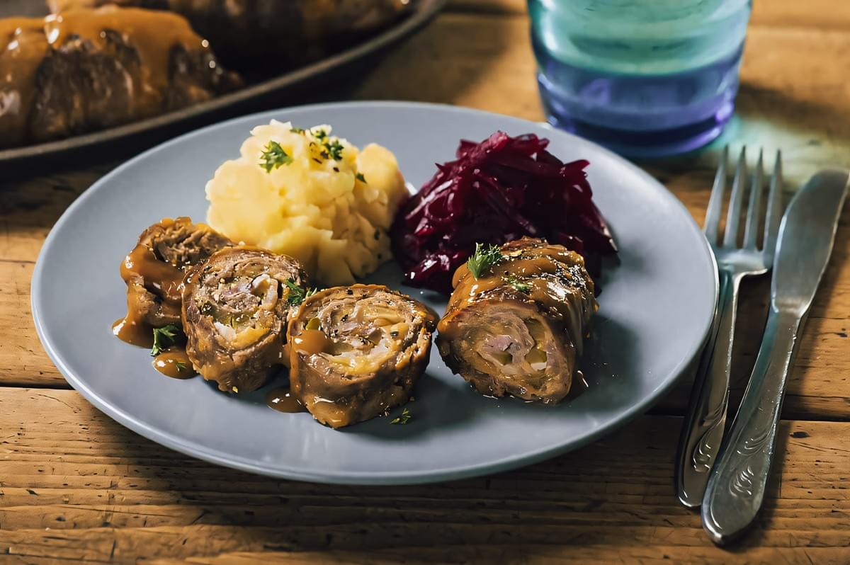 Close-up angled shot of traditional German rouladen with gravy, mashed potatoes, and red cabbage on ceramic plate, rustic dining setting highlighting hearty comfort food adapted for tropical kitchens Close-up angled shot of traditional German rouladen with gravy, mashed potatoes, and red cabbage on ceramic plate, rustic dining setting highlighting hearty comfort food adapted for tropical kitchens