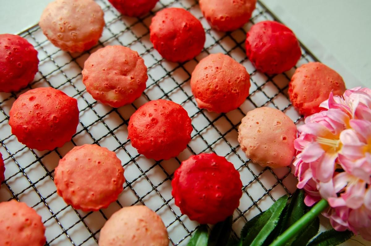 Rows of small, round baked goods in varying shades of pink and red are arranged on a black wire cooling rack. A cluster of pink artificial flowers rests in the bottom right corner, complementing the vibrant colors of the sweets. Rows of small, round baked goods in varying shades of pink and red are arranged on a black wire cooling rack. A cluster of pink artificial flowers rests in the bottom right corner, complementing the vibrant colors of the sweets.