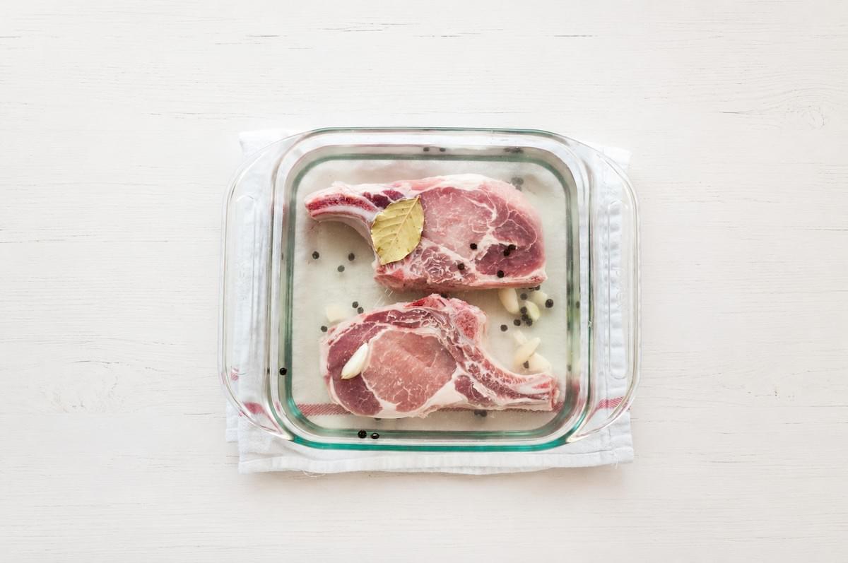 An overhead view of two raw, bone-in pork chops submerged in a clear liquid brine with a bay leaf, garlic cloves, and whole peppercorns inside a square glass dish, resting on a white surface. An overhead view of two raw, bone-in pork chops submerged in a clear liquid brine with a bay leaf, garlic cloves, and whole peppercorns inside a square glass dish, resting on a white surface.