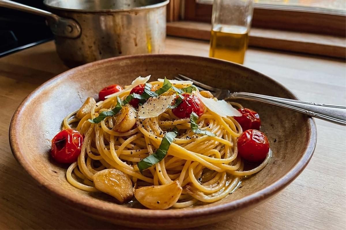 A bowl of spaghetti tossed with whole roasted garlic cloves, blistered cherry tomatoes, fresh basil ribbons, cracked black pepper, and shaved parmesan cheese, served on a wooden countertop next to a fork, olive oil bottle, and cooking pot. A bowl of spaghetti tossed with whole roasted garlic cloves, blistered cherry tomatoes, fresh basil ribbons, cracked black pepper, and shaved parmesan cheese, served on a wooden countertop next to a fork, olive oil bottle, and cooking pot.