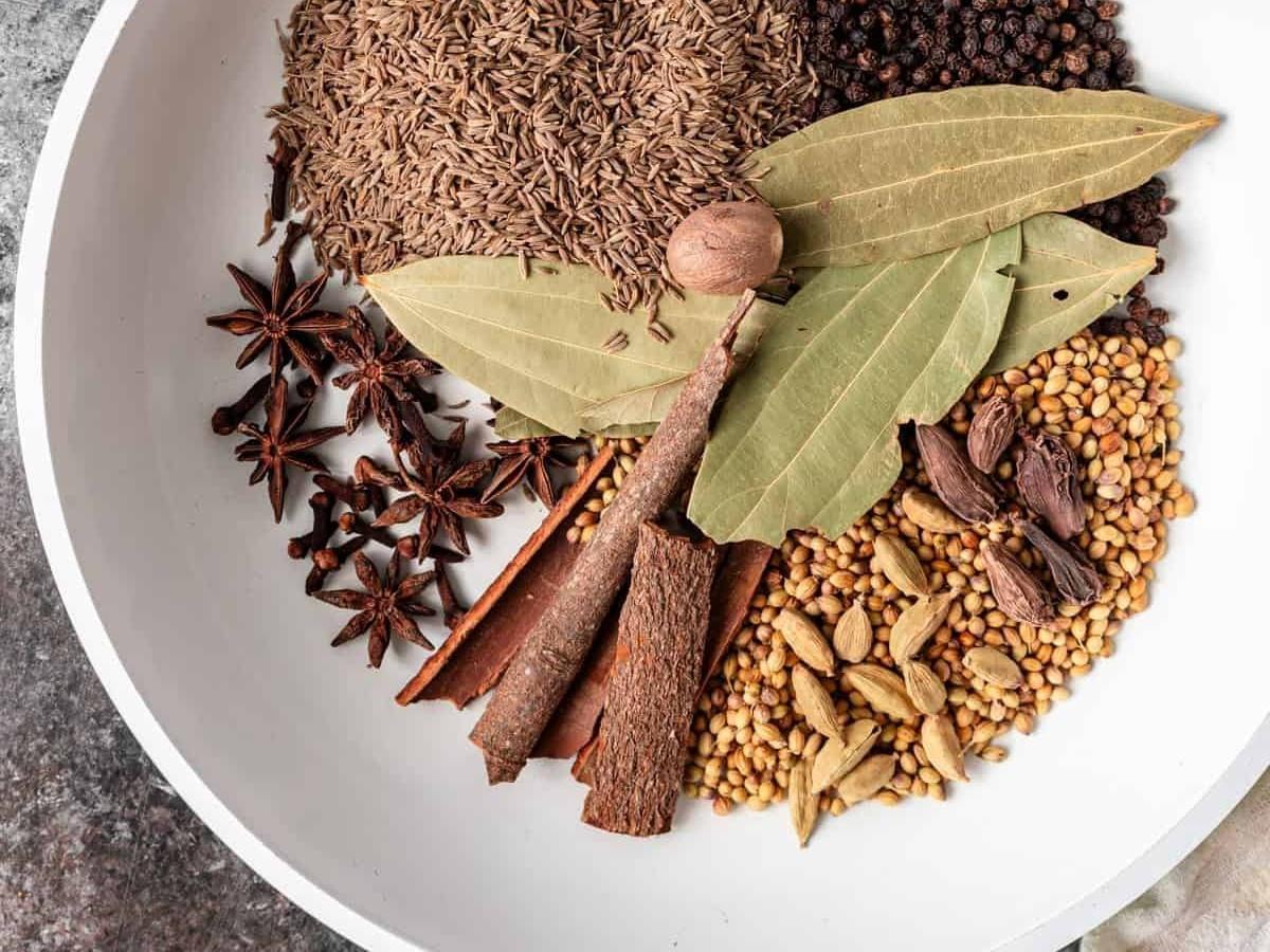 A white bowl holds an assortment of whole spices arranged in clusters, including cumin seeds, black peppercorns, coriander seeds, green and black cardamom pods, cinnamon sticks, star anise, cloves, bay leaves, and a single nutmeg. The textures and earthy tones of the spices contrast vividly against the smooth, light surface of the bowl. A white bowl holds an assortment of whole spices arranged in clusters, including cumin seeds, black peppercorns, coriander seeds, green and black cardamom pods, cinnamon sticks, star anise, cloves, bay leaves, and a single nutmeg. The textures and earthy tones of the spices contrast vividly against the smooth, light surface of the bowl.