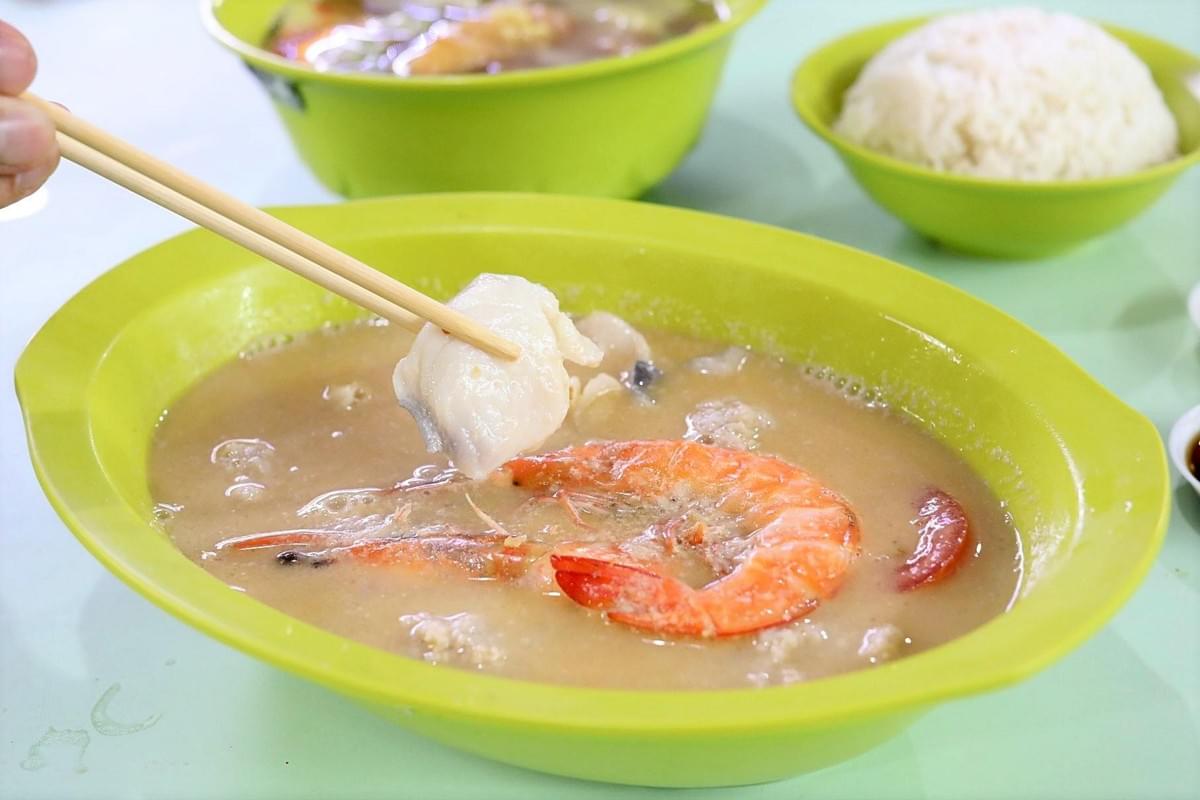 A pair of wooden chopsticks lifts a piece of white fish slice above a green bowl of seafood soup containing a large prawn. In the background sit another bowl of soup and a small green bowl filled with steamed white rice. A pair of wooden chopsticks lifts a piece of white fish slice above a green bowl of seafood soup containing a large prawn. In the background sit another bowl of soup and a small green bowl filled with steamed white rice.