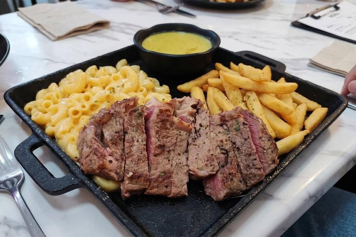 Slices of medium-rare steak served on a black cast iron skillet with macaroni and cheese, french fries, and a side of yellow sauce. Slices of medium-rare steak served on a black cast iron skillet with macaroni and cheese, french fries, and a side of yellow sauce.