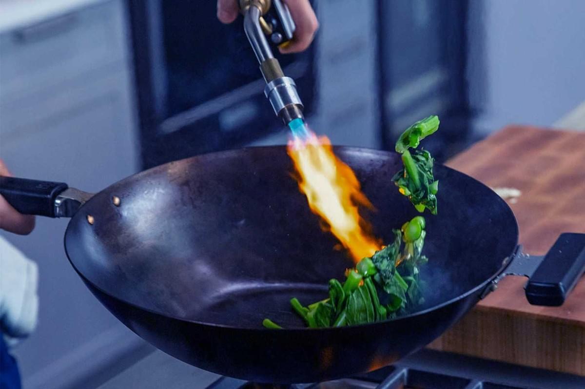 A person uses a blowtorch to cook leafy greens in a black wok, creating a vibrant flame. The scene conveys a dynamic and modern kitchen technique Wok Hei A person uses a blowtorch to cook leafy greens in a black wok, creating a vibrant flame. The scene conveys a dynamic and modern kitchen technique Wok Hei
