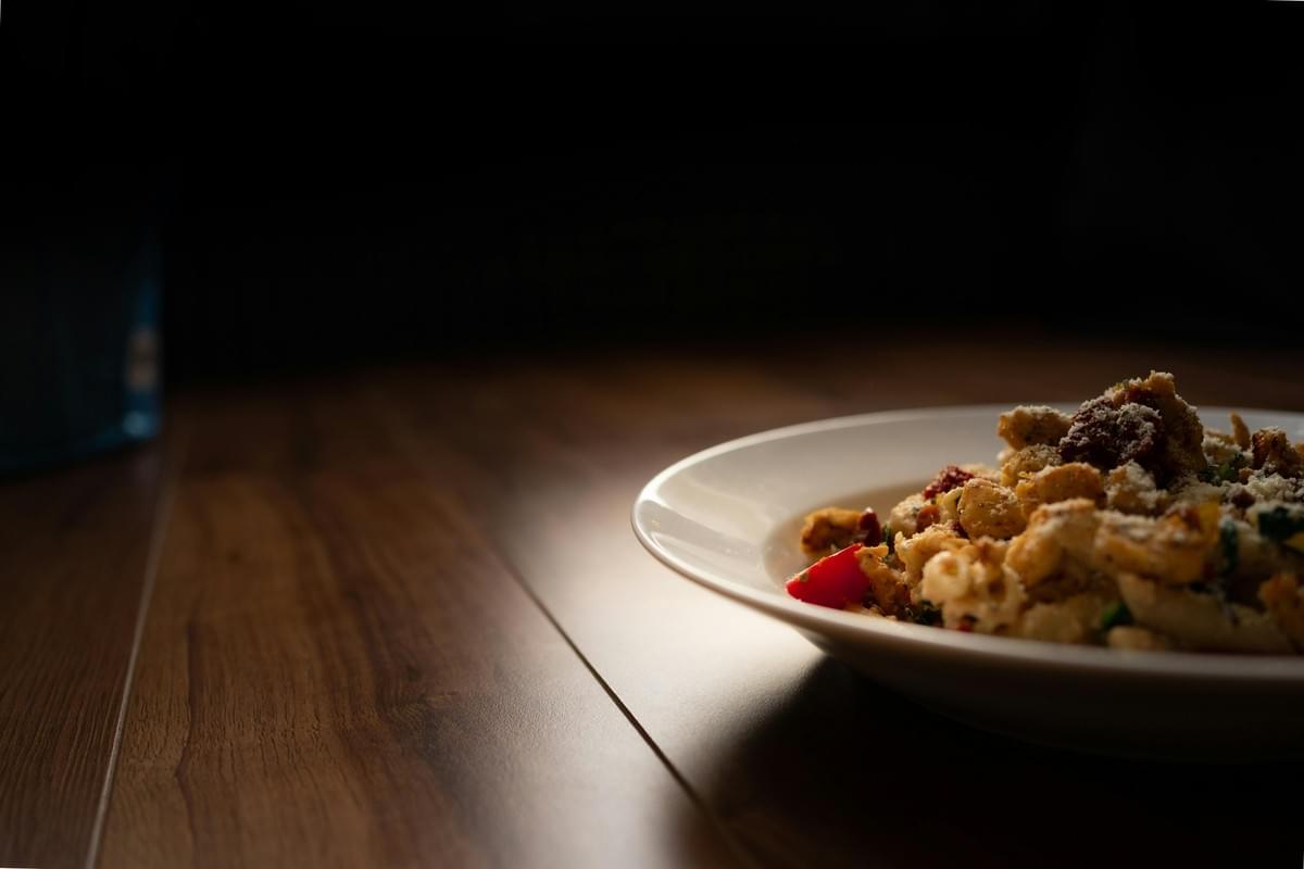 A shallow-focus, low-angle shot shows a white bowl filled with a pasta dish topped with grated cheese, sun-dried tomatoes, and fresh red bell peppers. The bowl sits on a polished dark wood table, dramatically lit from the side against a dark, shadowed background. A shallow-focus, low-angle shot shows a white bowl filled with a pasta dish topped with grated cheese, sun-dried tomatoes, and fresh red bell peppers. The bowl sits on a polished dark wood table, dramatically lit from the side against a dark, shadowed background.