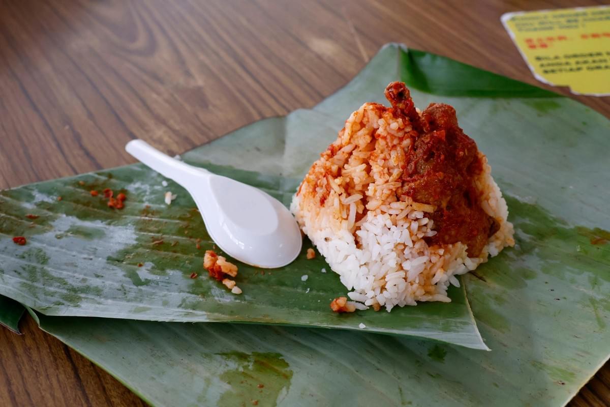 A pyramid-shaped mound of white and orange-tinted rice is topped with meat in a red chili sauce, served on a green banana leaf next to a white plastic spoon. The dish rests on a wooden table, capturing a casual, authentic Southeast Asian dining experience. A pyramid-shaped mound of white and orange-tinted rice is topped with meat in a red chili sauce, served on a green banana leaf next to a white plastic spoon. The dish rests on a wooden table, capturing a casual, authentic Southeast Asian dining experience.