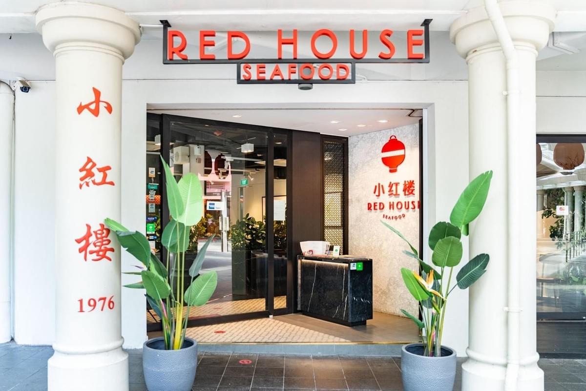 The entrance of Red House Seafood is shown, featuring red signage above glass doors and a reception desk inside. Potted green plants flank the doorway, set between white structural pillars with Chinese calligraphy. The entrance of Red House Seafood is shown, featuring red signage above glass doors and a reception desk inside. Potted green plants flank the doorway, set between white structural pillars with Chinese calligraphy.