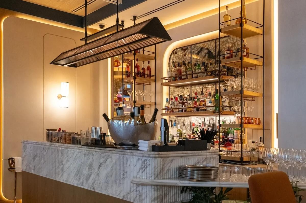 Maison Boulud restaurant bar area with a fluted marble counter, champagne chiller, and backlit liquor shelves. Maison Boulud restaurant bar area with a fluted marble counter, champagne chiller, and backlit liquor shelves.