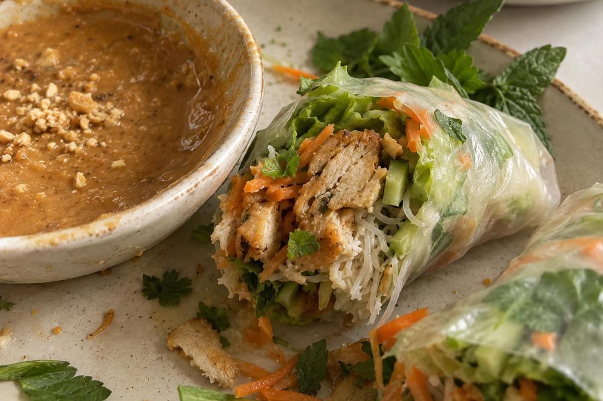 A close-up shot showcases a cut rice paper roll filled with seasoned chicken, vermicelli, shredded carrots, cucumber, and herbs resting on a ceramic platter next to a bowl of peanut-free satay sauce topped with crushed seeds. A close-up shot showcases a cut rice paper roll filled with seasoned chicken, vermicelli, shredded carrots, cucumber, and herbs resting on a ceramic platter next to a bowl of peanut-free satay sauce topped with crushed seeds.