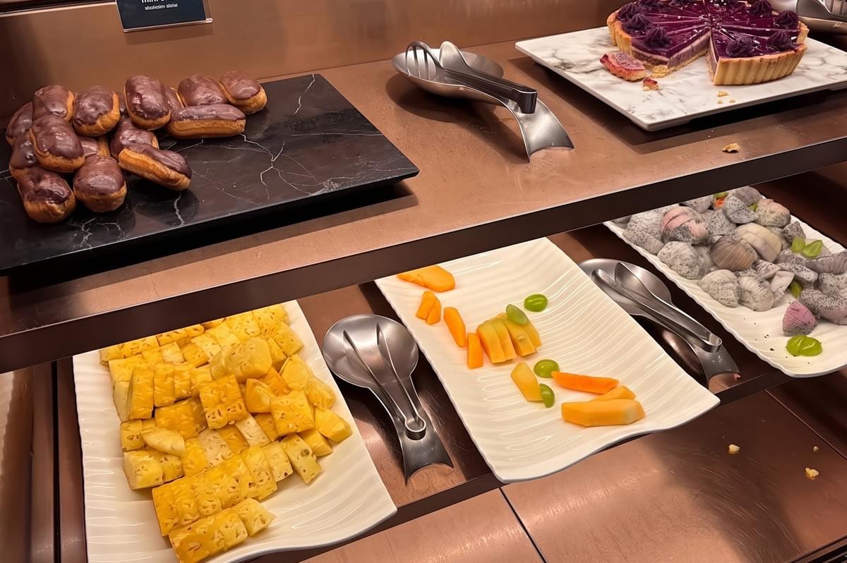A hotel buffet display featuring platters of chocolate eclairs, fresh pineapple, sliced cantaloupe and grapes, and dragon fruit pieces. A hotel buffet display featuring platters of chocolate eclairs, fresh pineapple, sliced cantaloupe and grapes, and dragon fruit pieces.