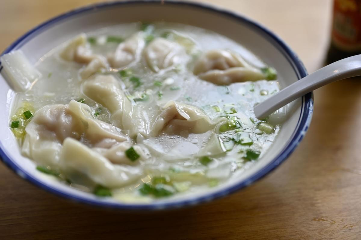 A blue-rimmed ceramic bowl sits on a wooden table filled with steaming clear broth and delicate wontons topped with chopped scallions. A white plastic soup spoon rests inside the bowl, resting against the side. A blue-rimmed ceramic bowl sits on a wooden table filled with steaming clear broth and delicate wontons topped with chopped scallions. A white plastic soup spoon rests inside the bowl, resting against the side.