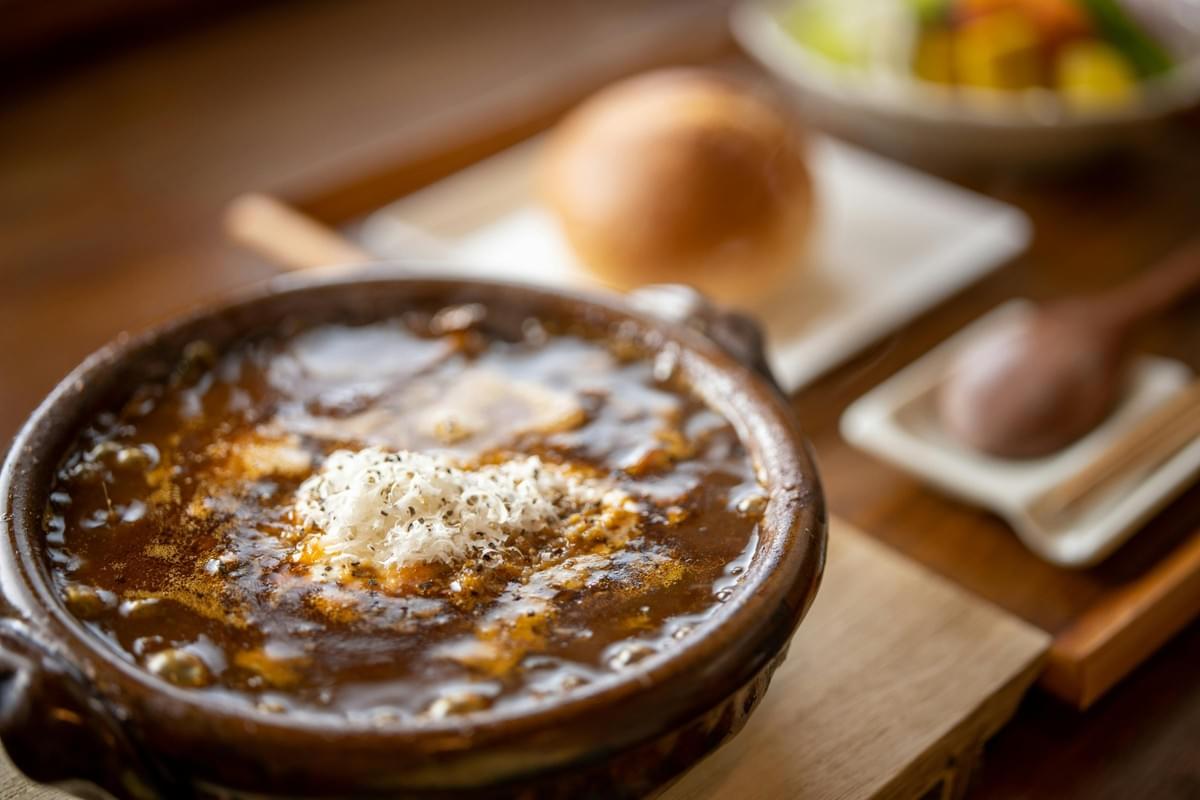 A earthenware bowl filled with rich, simmering dark stew garnished with shredded cheese and black pepper rests on a wooden tray in the foreground. In the soft-focused background, a bread roll and a side dish complement the warm dining arrangement. A earthenware bowl filled with rich, simmering dark stew garnished with shredded cheese and black pepper rests on a wooden tray in the foreground. In the soft-focused background, a bread roll and a side dish complement the warm dining arrangement.