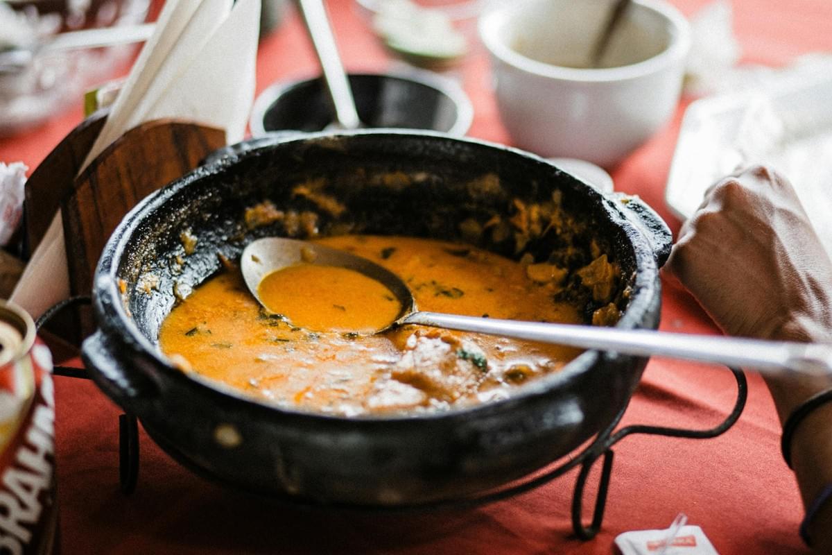 A traditional black clay pot sits on a metal stand, filled with a vibrant orange seafood stew and a large metal serving spoon. The dish is served on a red tablecloth surrounded by blurred dining elements, including a partial view of a person's arm and a beverage can. A traditional black clay pot sits on a metal stand, filled with a vibrant orange seafood stew and a large metal serving spoon. The dish is served on a red tablecloth surrounded by blurred dining elements, including a partial view of a person's arm and a beverage can.