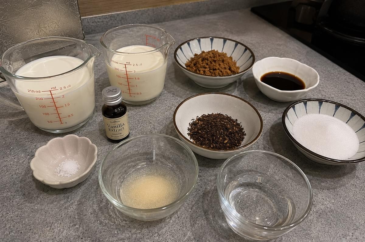 A flat-lay view of the ingredients required for the dessert, including two glass measuring cups of milk and cream, a bottle of vanilla extract, a small dish of salt, clear bowls of gelatin mixture and water, and small ceramic bowls filled with brown sugar, loose hojicha tea leaves, white sugar, and kuromitsu syrup. A flat-lay view of the ingredients required for the dessert, including two glass measuring cups of milk and cream, a bottle of vanilla extract, a small dish of salt, clear bowls of gelatin mixture and water, and small ceramic bowls filled with brown sugar, loose hojicha tea leaves, white sugar, and kuromitsu syrup.