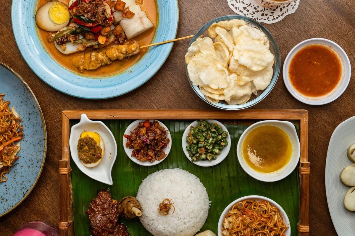 An overhead shot displays an assortment of traditional Indonesian dishes arranged neatly across a wooden tabletop. The centerpiece features a bamboo tray lined with a banana leaf, hosting a mound of white rice surrounded by small bowls of noodles, sambal, green beans, and seasoned egg. An overhead shot displays an assortment of traditional Indonesian dishes arranged neatly across a wooden tabletop. The centerpiece features a bamboo tray lined with a banana leaf, hosting a mound of white rice surrounded by small bowls of noodles, sambal, green beans, and seasoned egg.