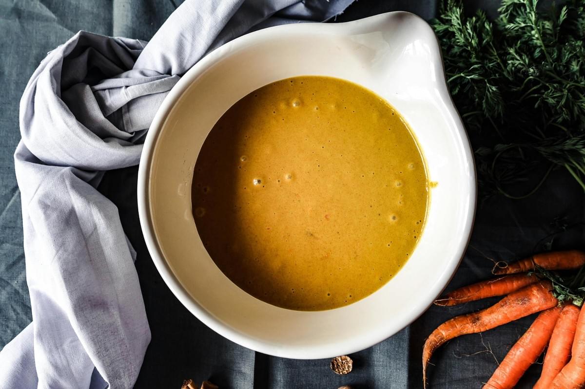 A large white ceramic bowl filled with creamy orange carrot soup sits at the center of the frame. It is set against a dark tablecloth with fresh whole carrots, greenery, spices, and a draped light blue cloth surrounding it. A large white ceramic bowl filled with creamy orange carrot soup sits at the center of the frame. It is set against a dark tablecloth with fresh whole carrots, greenery, spices, and a draped light blue cloth surrounding it.