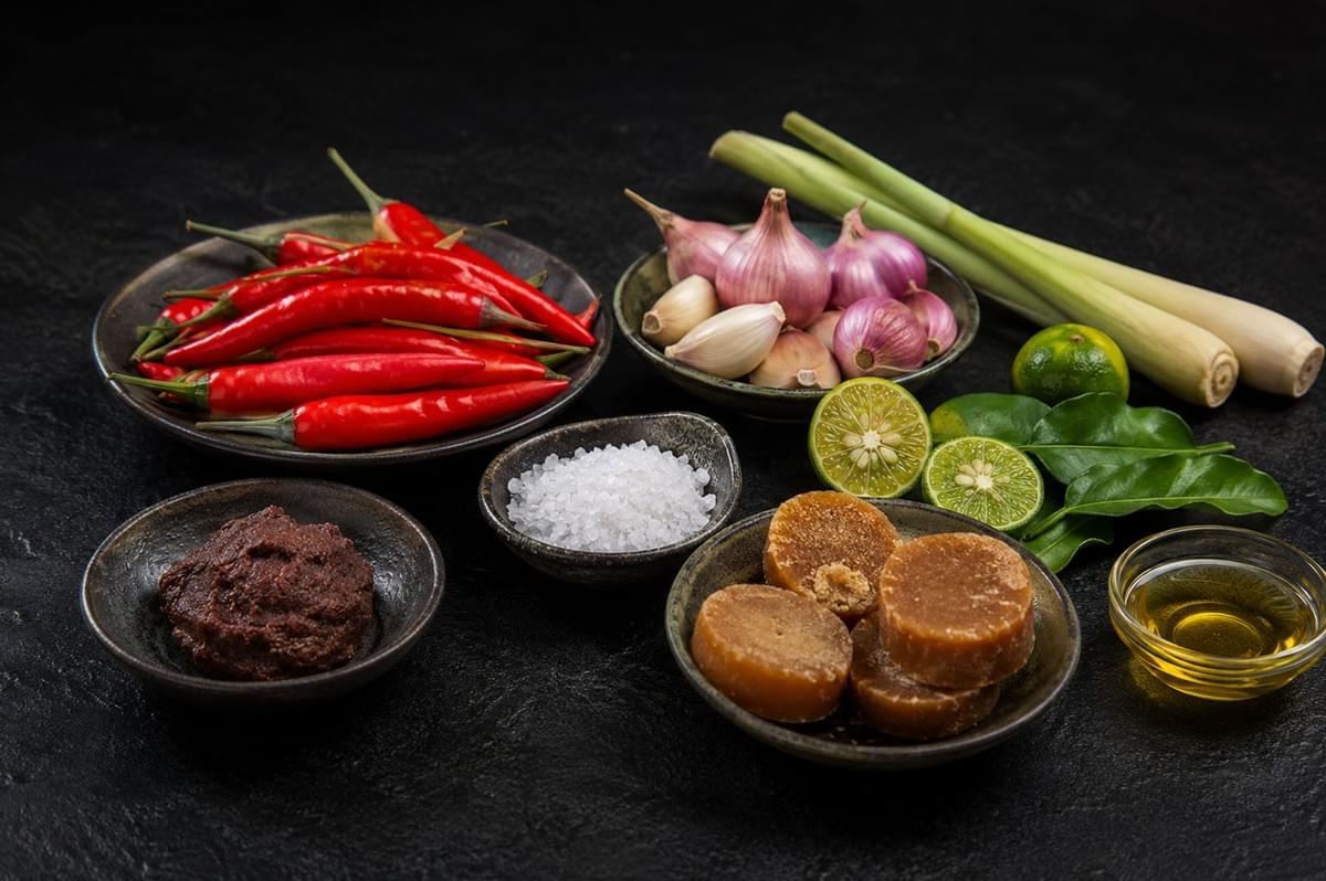 Fresh ingredients for sambal including red chilies, shallots, garlic, lemongrass, lime leaves, palm sugar, and shrimp paste arranged on black surface Fresh ingredients for sambal including red chilies, shallots, garlic, lemongrass, lime leaves, palm sugar, and shrimp paste arranged on black surface