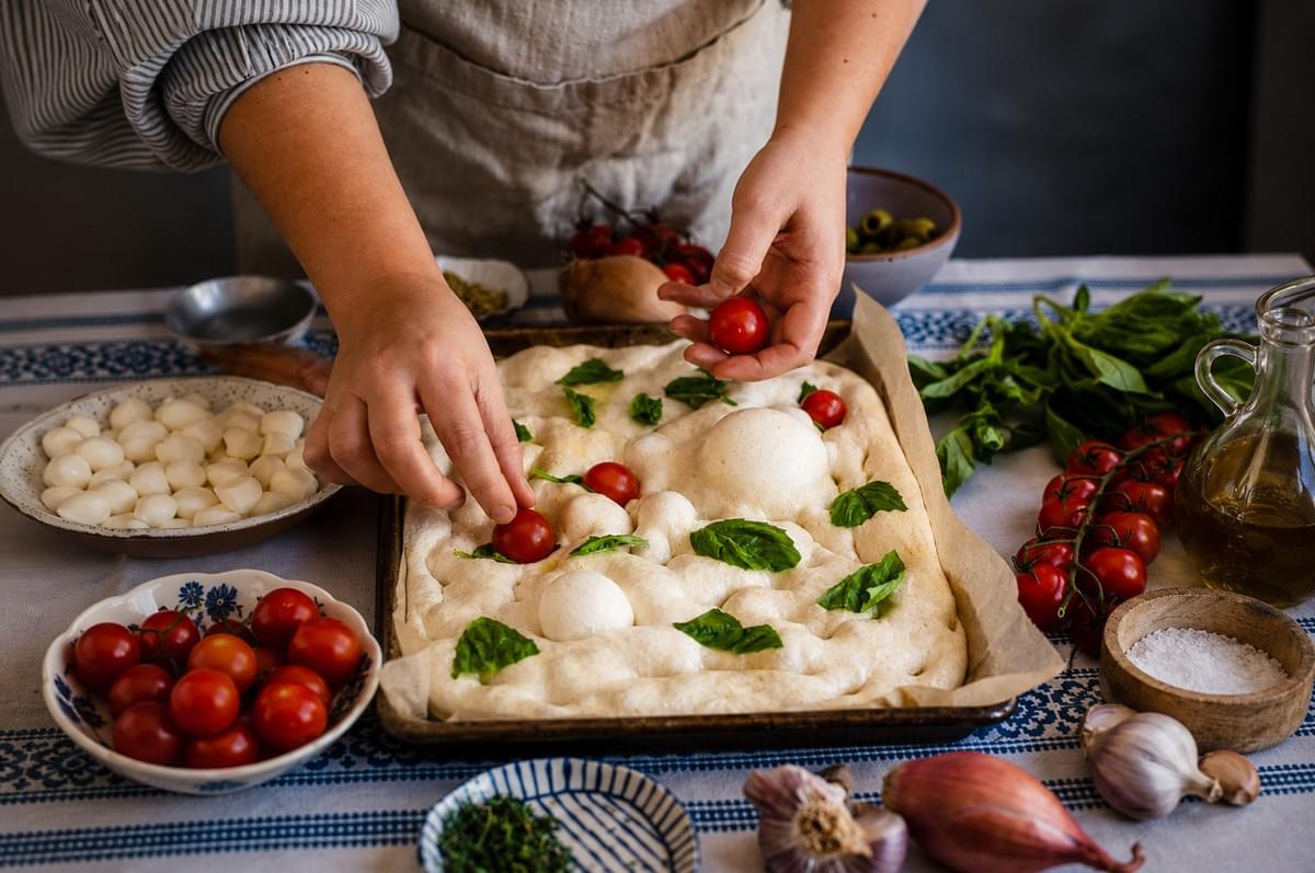 A person in an apron placing cherry tomatoes and fresh basil leaves onto unbaked focaccia dough in a baking pan, surrounded by bowls of mozzarella, tomatoes, and other fresh ingredients on a patterned tablecloth. A person in an apron placing cherry tomatoes and fresh basil leaves onto unbaked focaccia dough in a baking pan, surrounded by bowls of mozzarella, tomatoes, and other fresh ingredients on a patterned tablecloth.