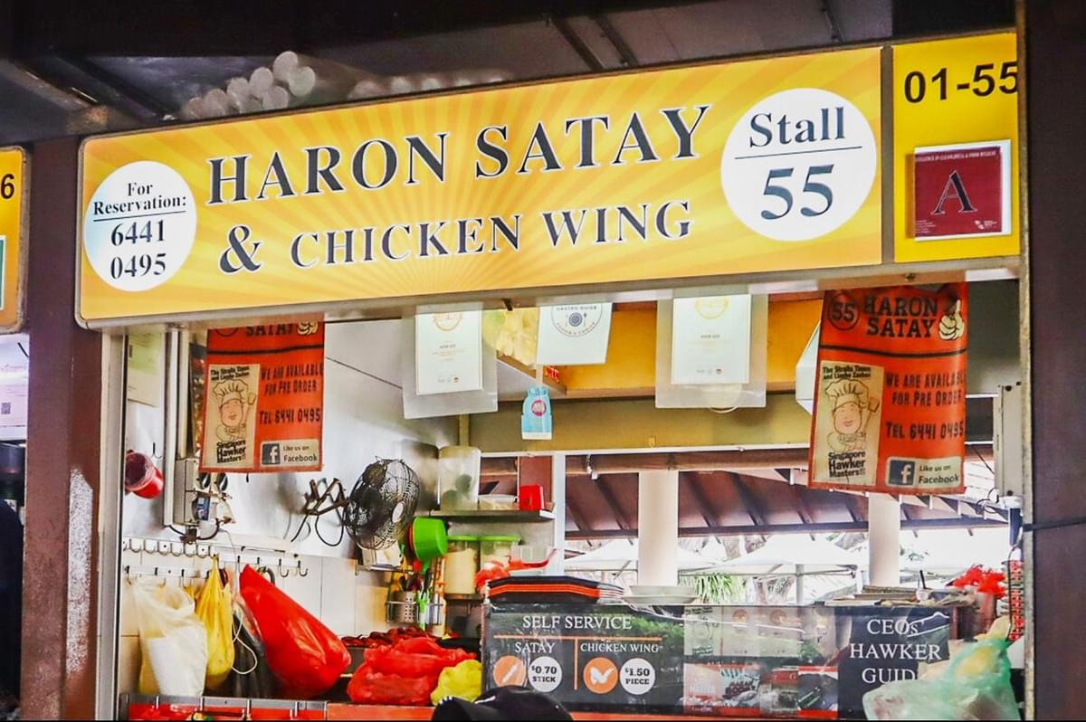 Food stall with a bright yellow sign reading "Haron Satay & Chicken Wing, Stall 55". The counter displays menu signs and reservation contact details, evoking a lively hawker atmosphere. Food stall with a bright yellow sign reading "Haron Satay & Chicken Wing, Stall 55". The counter displays menu signs and reservation contact details, evoking a lively hawker atmosphere.
