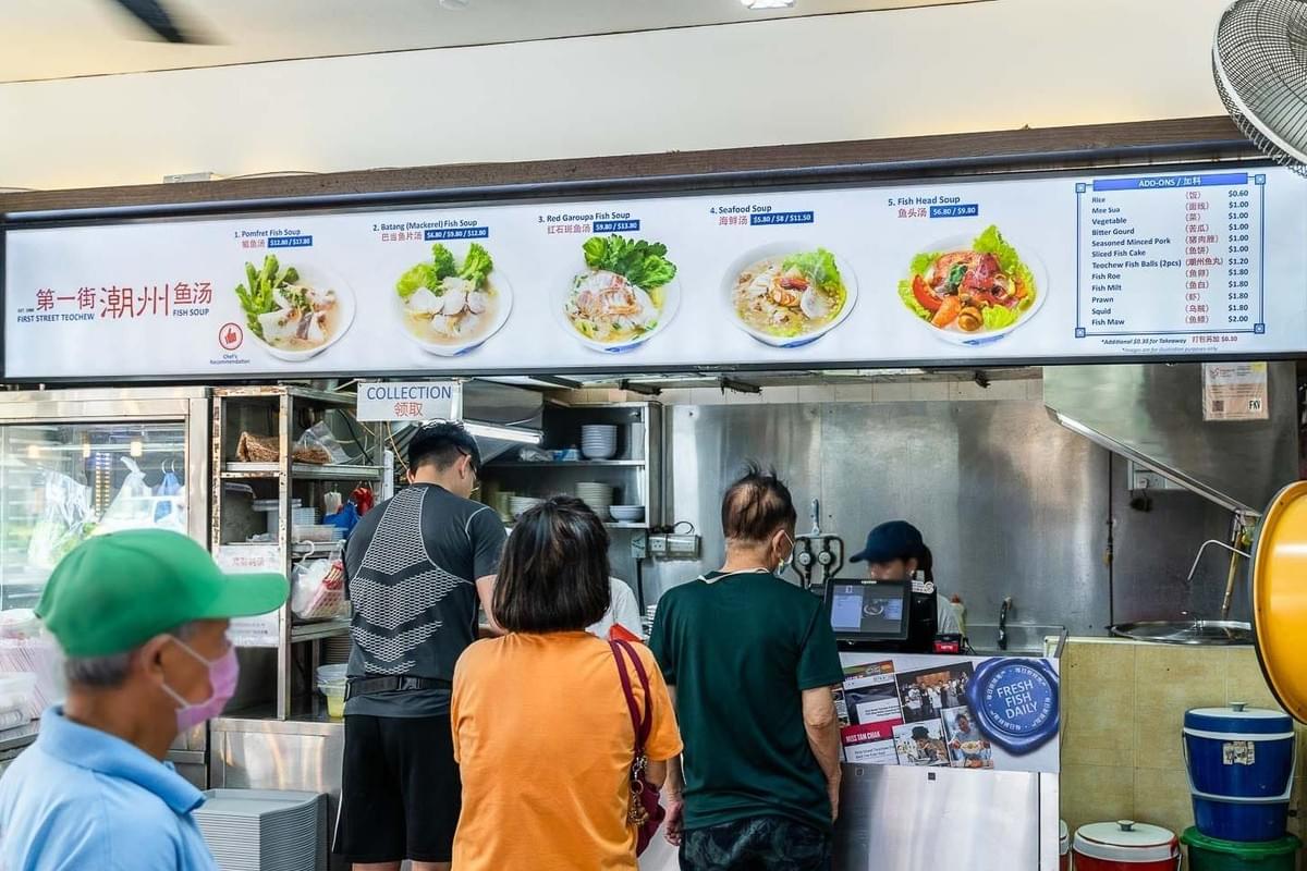 Customers queue in front of a food stall counter for First Street Teochew Fish Soup. Overhead, a large illuminated menu displays photos of various fish soup dishes along with prices and add-on options. Customers queue in front of a food stall counter for First Street Teochew Fish Soup. Overhead, a large illuminated menu displays photos of various fish soup dishes along with prices and add-on options.