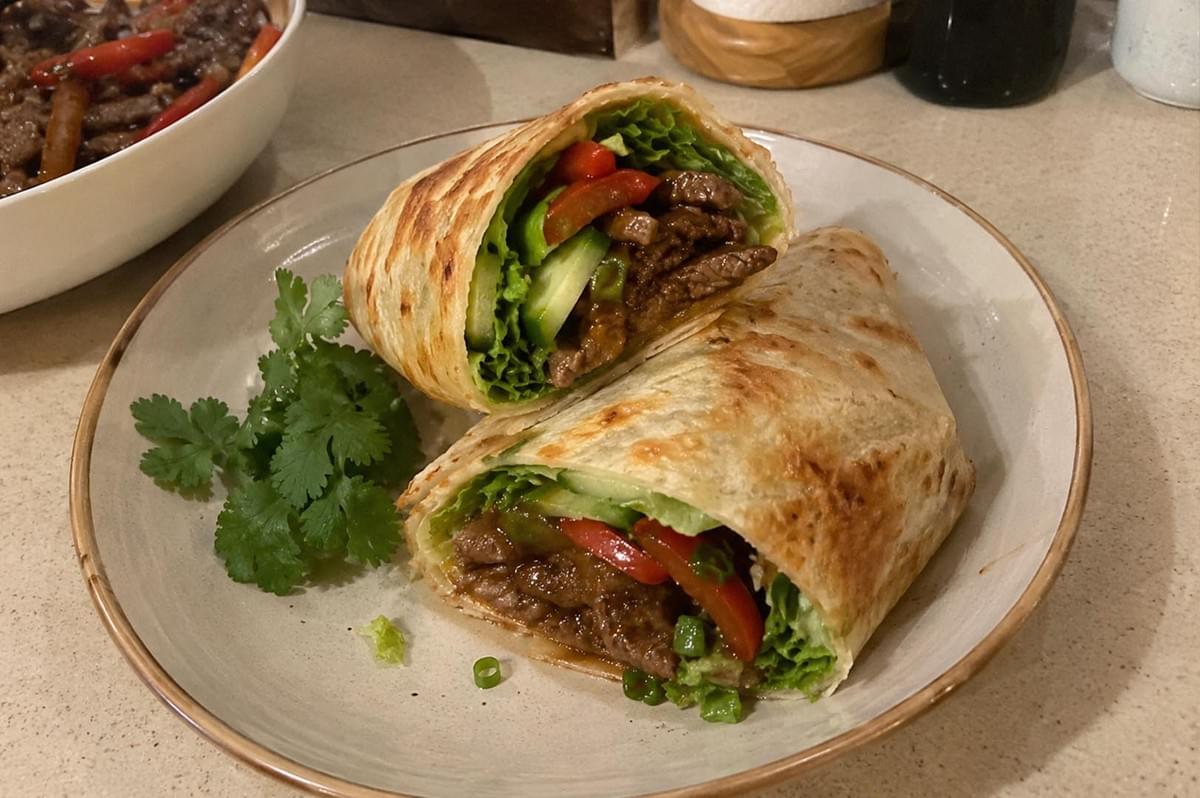 A close-up shot of a black pepper beef roti wrap cut diagonally into two halves, resting on a beige ceramic plate alongside fresh cilantro leaves. The golden-brown, flaky roti flatbread is tightly rolled and filled with savory stir-fried beef slices, crisp green lettuce, sliced red bell peppers, and fresh cucumber spears. The plate sits on a neutral countertop with a bowl of extra beef stir-fry visible in the soft-focus background. A close-up shot of a black pepper beef roti wrap cut diagonally into two halves, resting on a beige ceramic plate alongside fresh cilantro leaves. The golden-brown, flaky roti flatbread is tightly rolled and filled with savory stir-fried beef slices, crisp green lettuce, sliced red bell peppers, and fresh cucumber spears. The plate sits on a neutral countertop with a bowl of extra beef stir-fry visible in the soft-focus background.