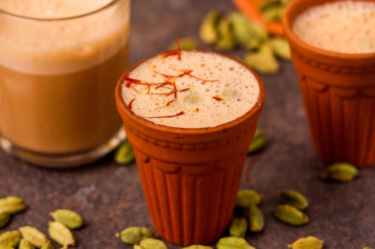 Frothy saffron cardamom tea served in traditional clay cups (kulhad) with whole green cardamom pods scattered on a dark surface. Frothy saffron cardamom tea served in traditional clay cups (kulhad) with whole green cardamom pods scattered on a dark surface.