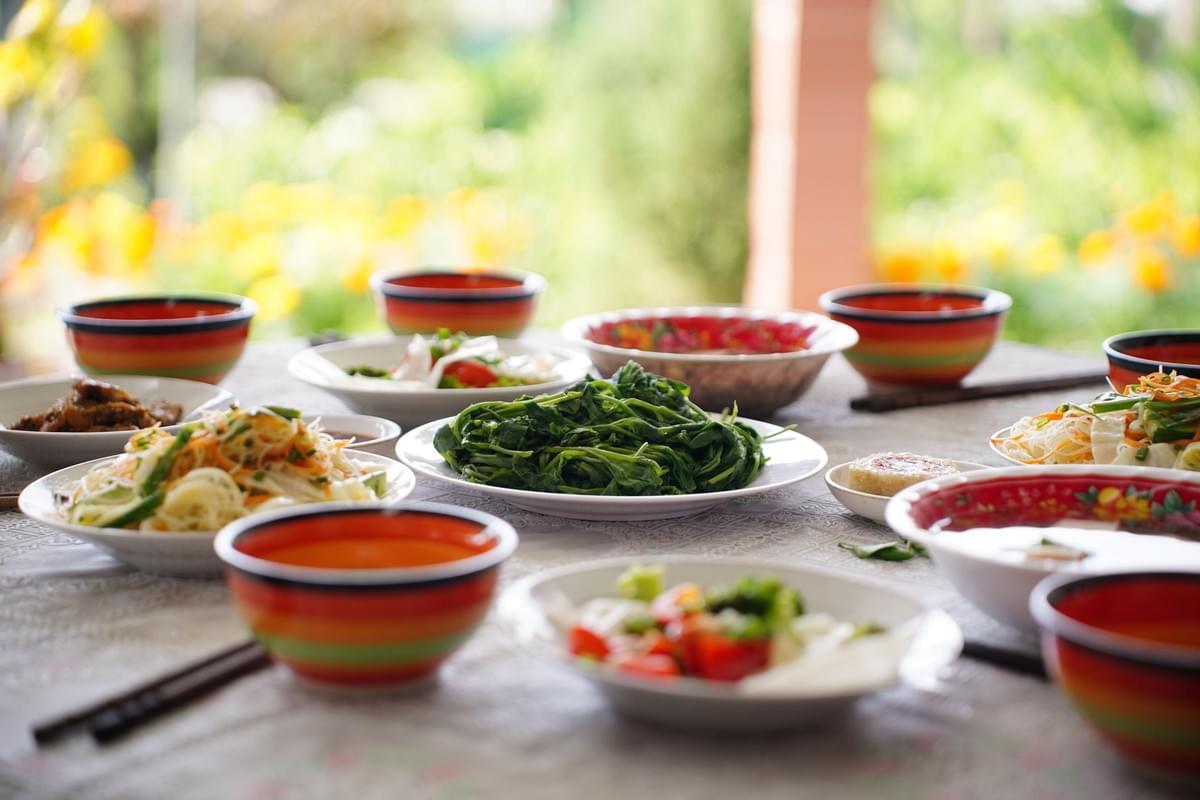 A dining table is spread with a variety of colorful dishes, including leafy greens, salads, and soups served in matching striped bowls. The meal is set outdoors on a sunlit patio overlooking a lush, blurred green garden. A dining table is spread with a variety of colorful dishes, including leafy greens, salads, and soups served in matching striped bowls. The meal is set outdoors on a sunlit patio overlooking a lush, blurred green garden.