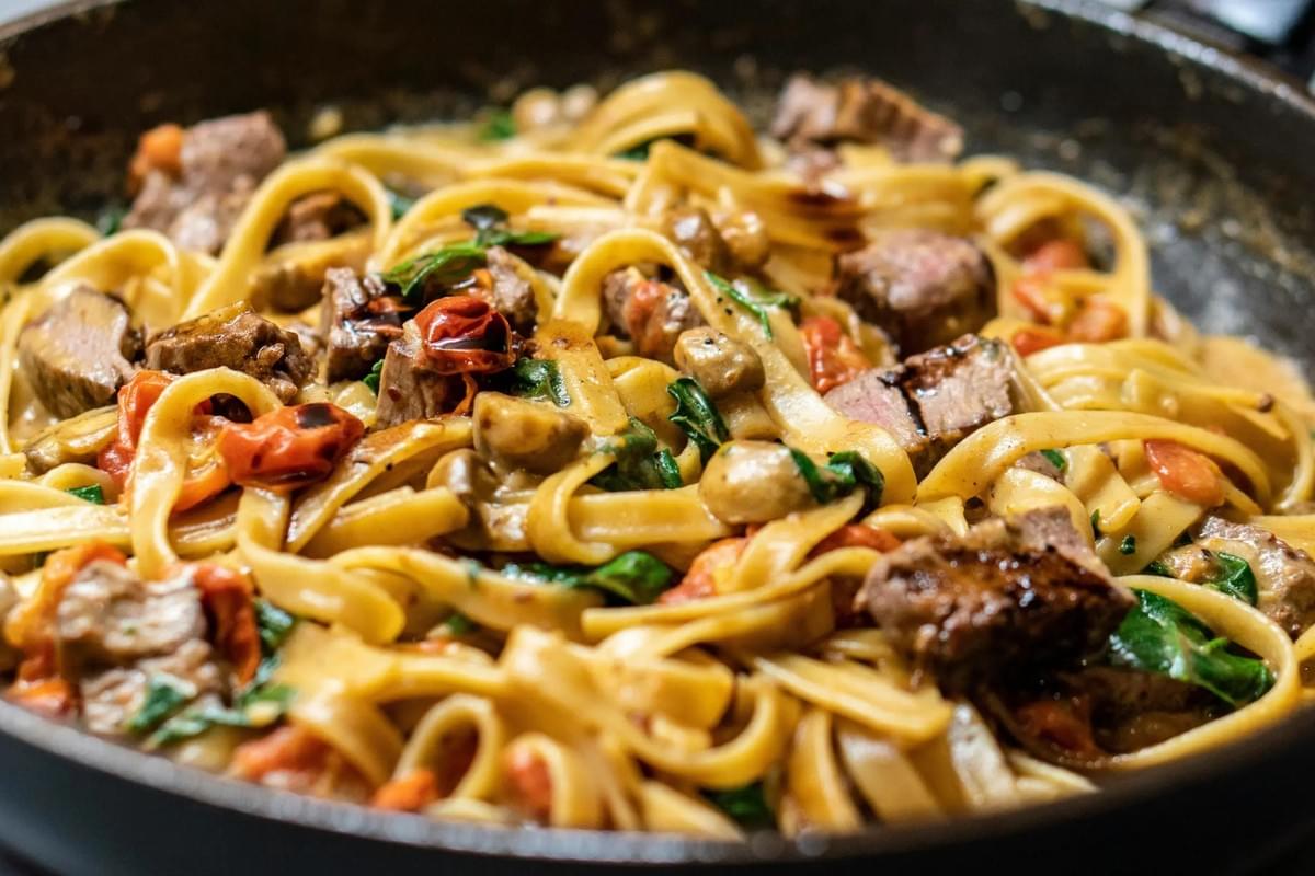 A skillet is filled with a creamy pasta dish featuring long, flat noodles. The meal is tossed with seared steak bites, sautéed mushrooms, wilted greens, and burst cherry tomatoes. A skillet is filled with a creamy pasta dish featuring long, flat noodles. The meal is tossed with seared steak bites, sautéed mushrooms, wilted greens, and burst cherry tomatoes.