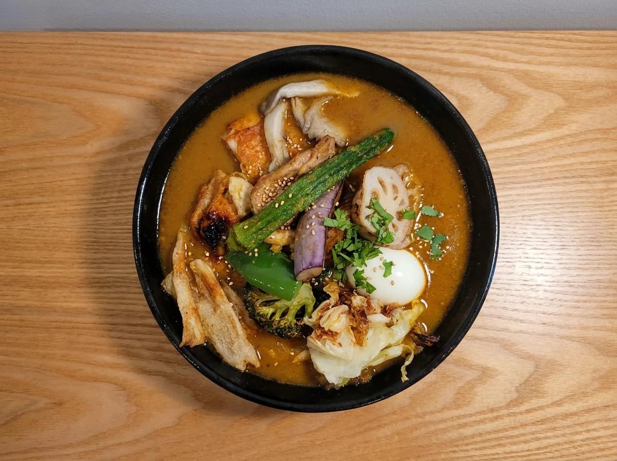 Top-down view of a rich Japanese soup curry with assorted vegetables, chicken, and a soft-boiled egg in a black bowl. Top-down view of a rich Japanese soup curry with assorted vegetables, chicken, and a soft-boiled egg in a black bowl.