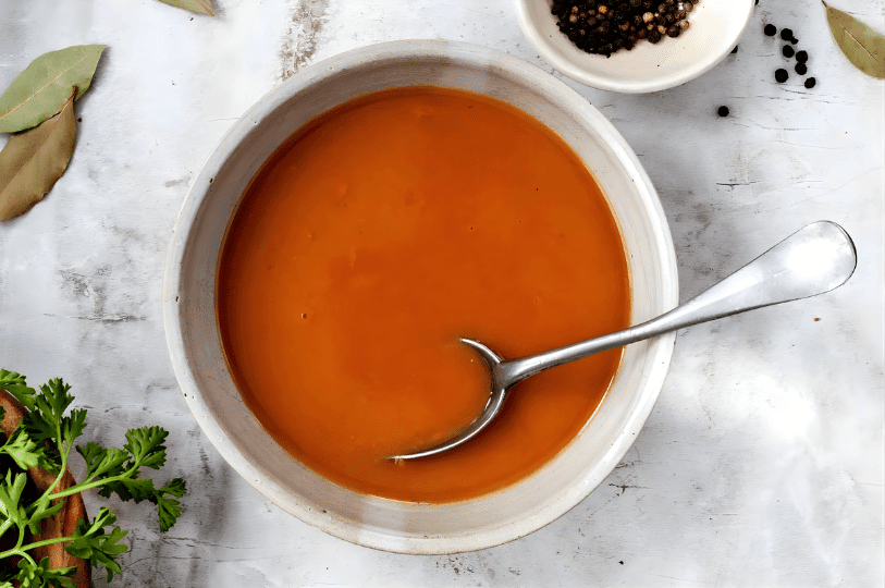 A top-down view of a bowl containing a rich, brown Espagnole sauce with a silver spoon resting inside, surrounded by dried bay leaves and black peppercorns on a marble surface. A top-down view of a bowl containing a rich, brown Espagnole sauce with a silver spoon resting inside, surrounded by dried bay leaves and black peppercorns on a marble surface.