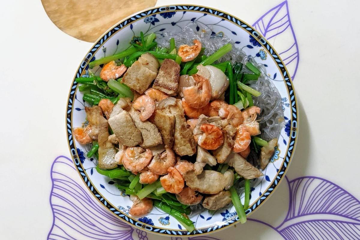 A vibrant bowl of glass noodles topped with cooked small shrimp, sliced pork, and fresh green leafy vegetables sits on a patterned surface. The dish is presented in a patterned ceramic bowl with a floral rim design. A vibrant bowl of glass noodles topped with cooked small shrimp, sliced pork, and fresh green leafy vegetables sits on a patterned surface. The dish is presented in a patterned ceramic bowl with a floral rim design.
