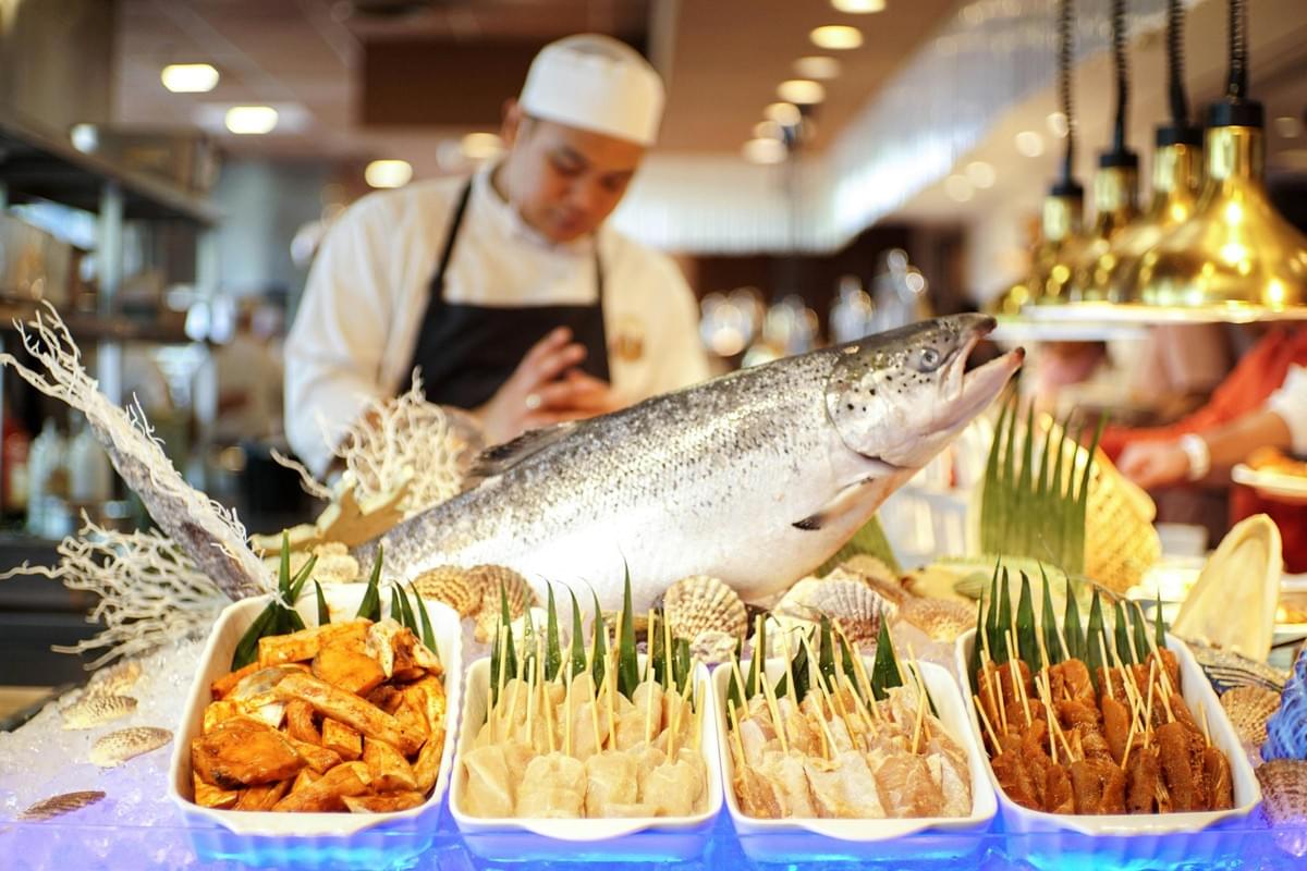 A large whole salmon and several trays of skewers sit on ice at a brightly lit seafood buffet display. In the softly blurred background, a chef wearing a white uniform and hat stands behind the food counter. A large whole salmon and several trays of skewers sit on ice at a brightly lit seafood buffet display. In the softly blurred background, a chef wearing a white uniform and hat stands behind the food counter.