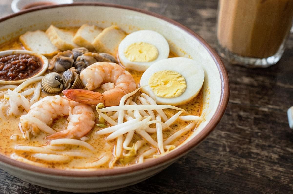 Close-up of a bowl of creamy laksa noodles topped with shrimp, mussels, bean sprouts, fish cake, sliced hard-boiled eggs, and sambal. Close-up of a bowl of creamy laksa noodles topped with shrimp, mussels, bean sprouts, fish cake, sliced hard-boiled eggs, and sambal.
