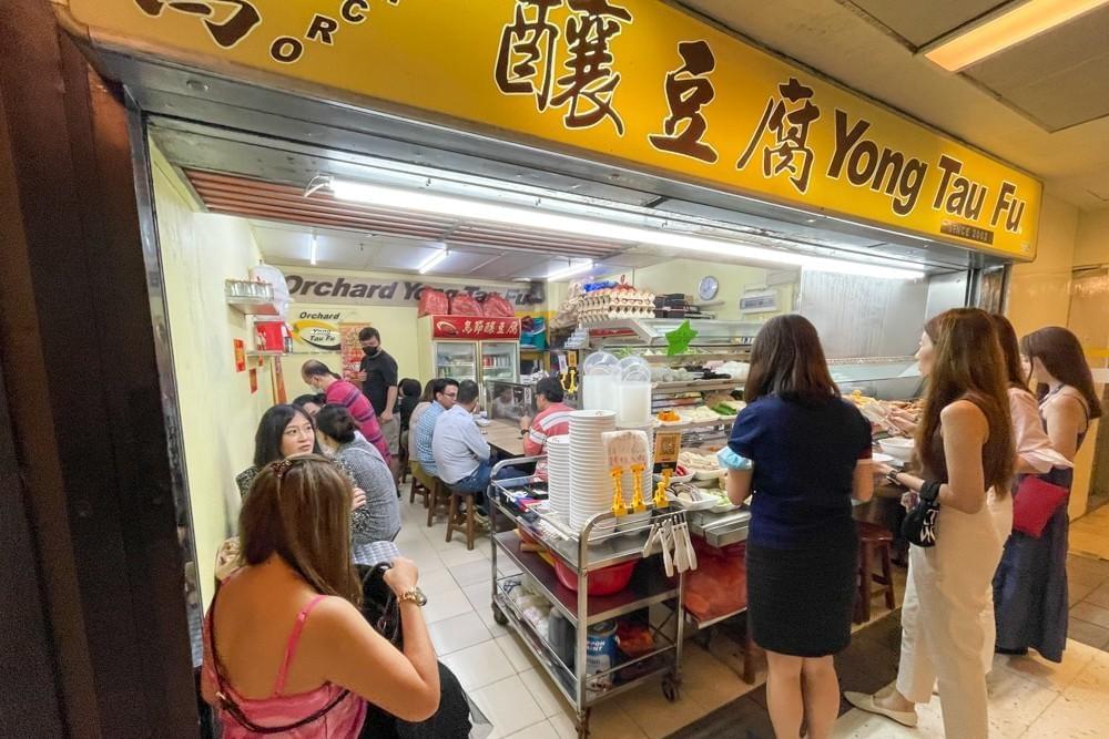 A brightly lit, narrow food stall named "Orchard Yong Tau Fu" features a vibrant yellow sign and a line of customers waiting to order. Inside, patrons sit at compact wooden tables and stools while staff prepare ingredients displayed on a metal service cart. A brightly lit, narrow food stall named "Orchard Yong Tau Fu" features a vibrant yellow sign and a line of customers waiting to order. Inside, patrons sit at compact wooden tables and stools while staff prepare ingredients displayed on a metal service cart.