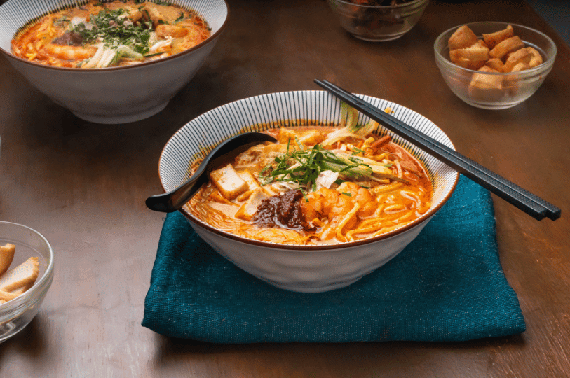 A steaming bowl of Singaporean laksa with thick noodles, shrimp, sliced fish cake, tofu puffs, and a dollop of sambal chili paste, resting on a blue napkin with black chopsticks. A steaming bowl of Singaporean laksa with thick noodles, shrimp, sliced fish cake, tofu puffs, and a dollop of sambal chili paste, resting on a blue napkin with black chopsticks.