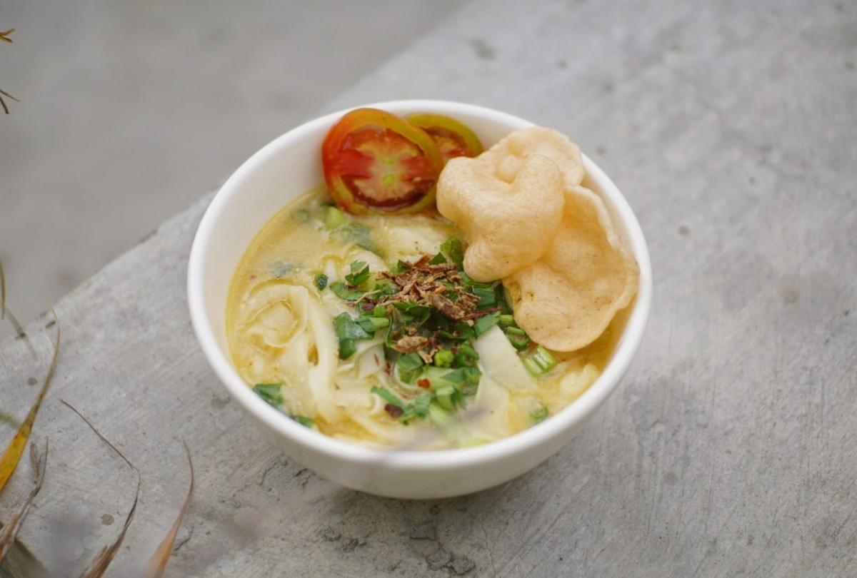 A white bowl filled with noodle soup topped with fresh herbs, fried shallots, sliced tomatoes, and crisp crackers. The bowl rests on a neutral concrete surface outdoors. A white bowl filled with noodle soup topped with fresh herbs, fried shallots, sliced tomatoes, and crisp crackers. The bowl rests on a neutral concrete surface outdoors.