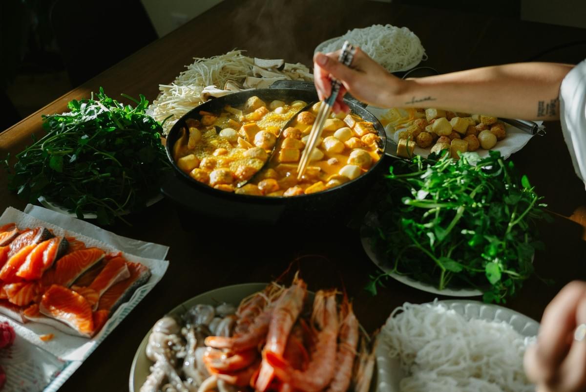 A bubbling hot pot sits at the center of a wooden table, filled with fish balls and tofu as a diner reaches in with chopsticks. Surrounding the broth is a generous spread of fresh ingredients prepared for dipping, including sliced salmon, prawns, enoki mushrooms, and leafy greens. A bubbling hot pot sits at the center of a wooden table, filled with fish balls and tofu as a diner reaches in with chopsticks. Surrounding the broth is a generous spread of fresh ingredients prepared for dipping, including sliced salmon, prawns, enoki mushrooms, and leafy greens.