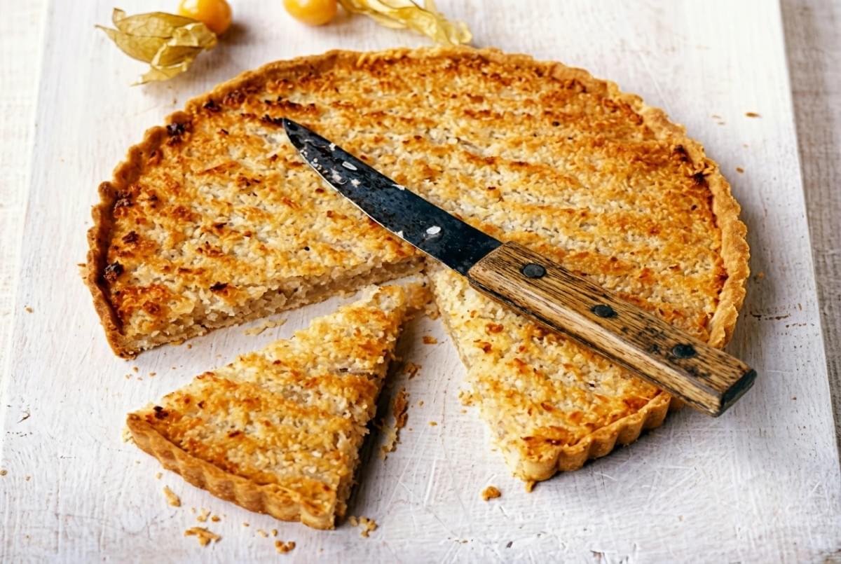 Baked coconut tart on a white wooden board with a slice cut out and a knife lying on top. Baked coconut tart on a white wooden board with a slice cut out and a knife lying on top.