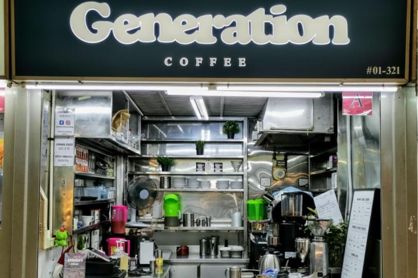 The storefront of "Generation Coffee," a modern hawker stall with a black sign, stainless steel counters, and various coffee-making equipment visible inside. The storefront of "Generation Coffee," a modern hawker stall with a black sign, stainless steel counters, and various coffee-making equipment visible inside.