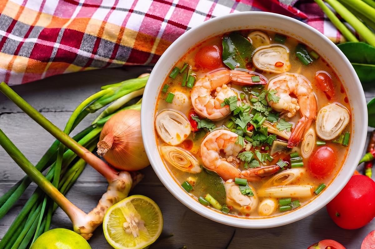 A bowl of authentic spicy Thai tom yum soup with shrimp, mushrooms, and fresh herbs surrounded by ingredients like lemongrass, tomatoes, and lime. A bowl of authentic spicy Thai tom yum soup with shrimp, mushrooms, and fresh herbs surrounded by ingredients like lemongrass, tomatoes, and lime.