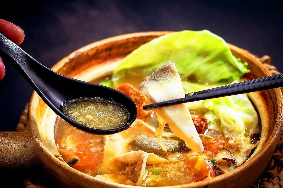 A clay pot holds hot soup filled with cabbage, mushrooms, and various meats. A black spoon scoops up savory broth while chopsticks hold a tender piece of fish skin above the dish. A clay pot holds hot soup filled with cabbage, mushrooms, and various meats. A black spoon scoops up savory broth while chopsticks hold a tender piece of fish skin above the dish.