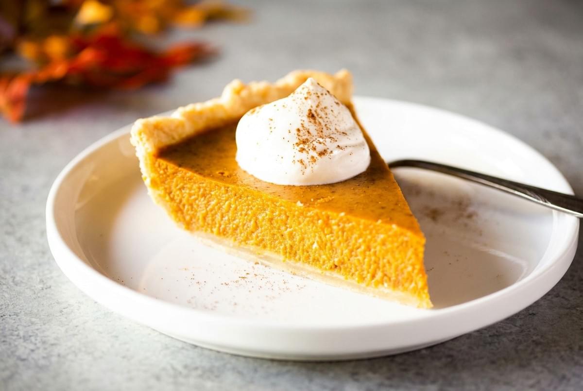 Slice of pumpkin pie topped with whipped cream and cinnamon on a white plate. Slice of pumpkin pie topped with whipped cream and cinnamon on a white plate.