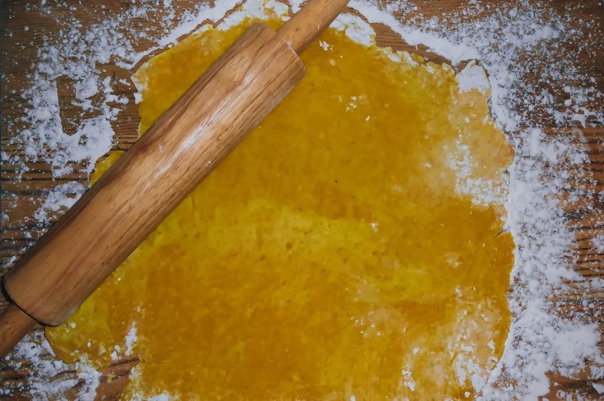 Top-down view of curry-infused Jamaican patty dough rolled flat with a wooden rolling pin on a floured surface. Top-down view of curry-infused Jamaican patty dough rolled flat with a wooden rolling pin on a floured surface.